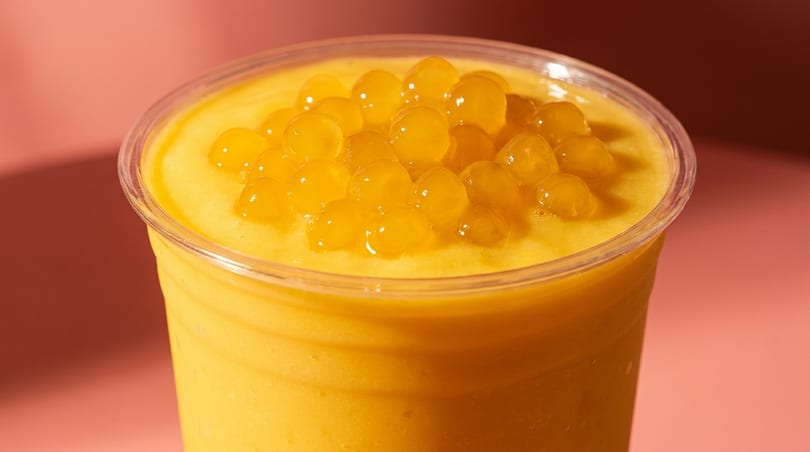 Close-up of a mango smoothie in a clear plastic cup, topped with golden popping boba.