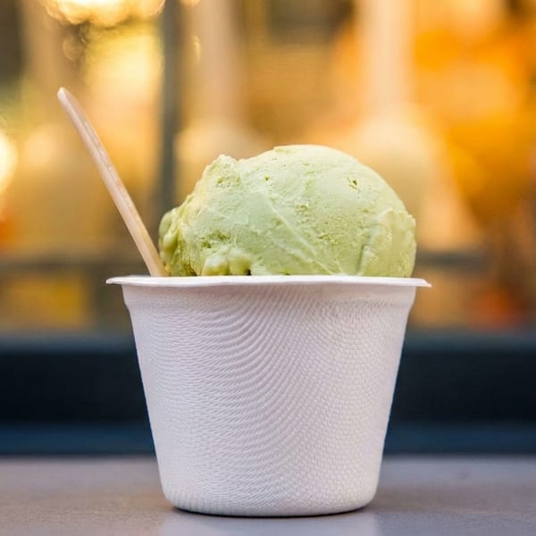 A scoop of green ice cream in a white cup with a wooden spoon, against a blurred background.
