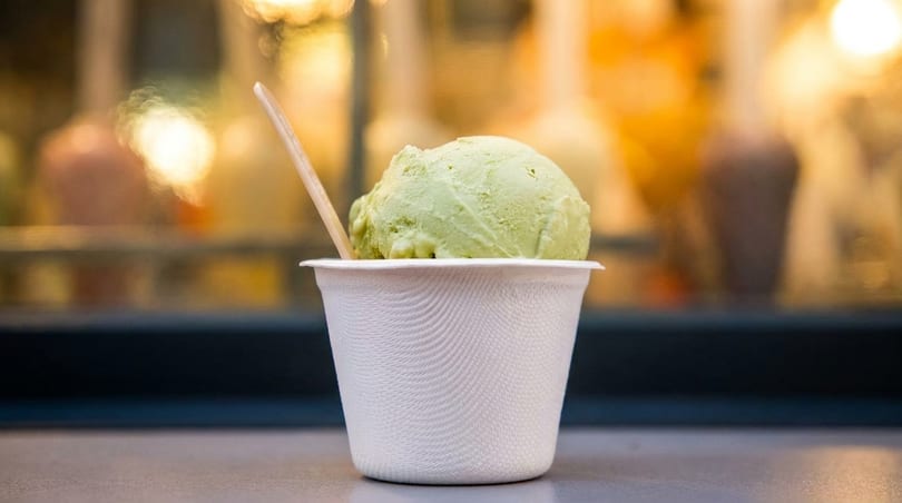 A scoop of green ice cream in a white cup with a wooden spoon, against a blurred background.